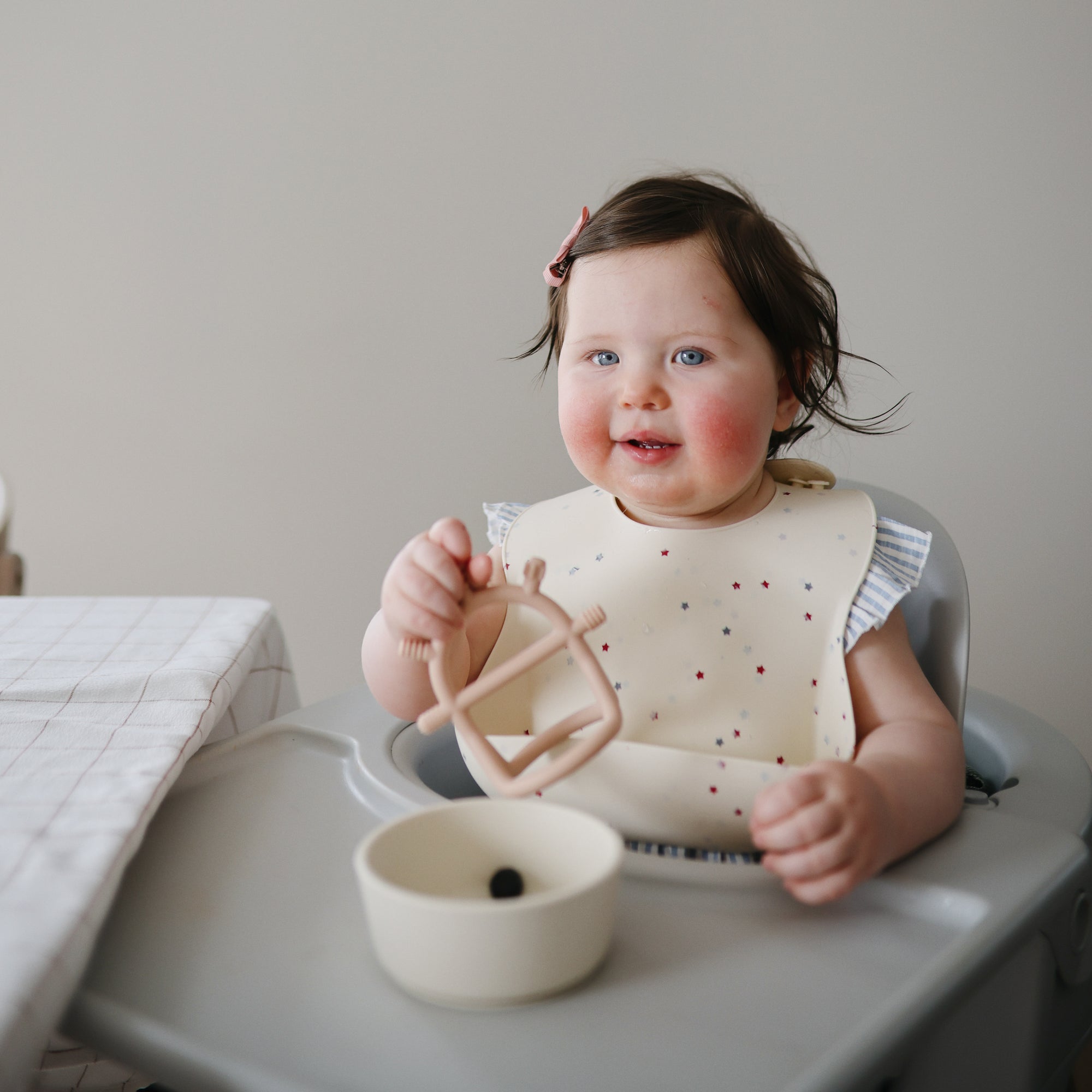 Smiling baby in high chair holds mushie No-Drop Sensory Teether, bowl on table nearby.
