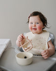 Smiling baby in high chair holds mushie No-Drop Sensory Teether, bowl on table nearby.