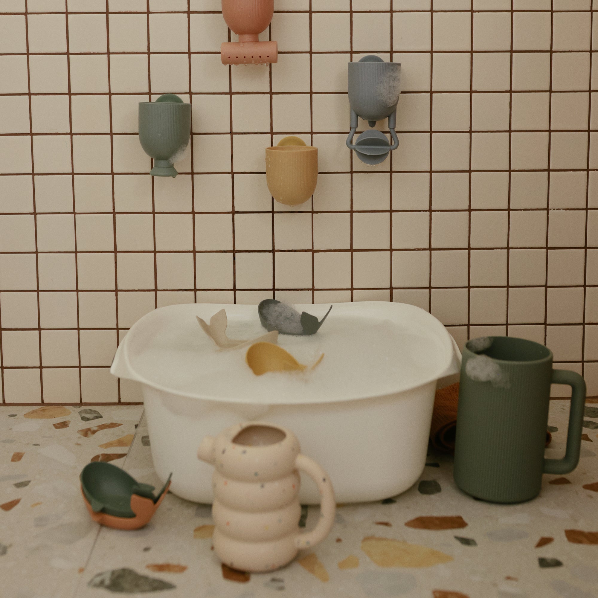 A white tub with Mushie Whale Bath Stackers, toy cups, and a green mug on tiled surfaces.