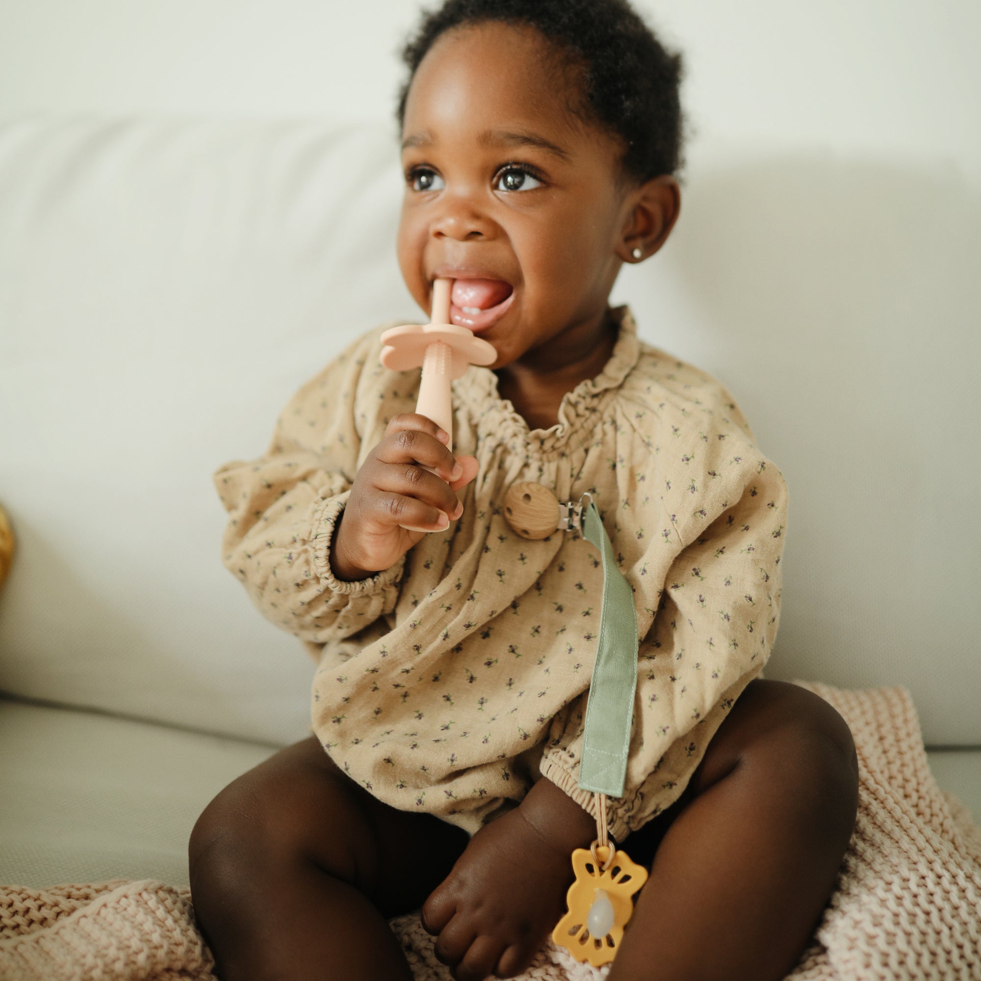A young child with curly hair smiles on a couch, wearing a beige patterned outfit and holding a pink silicone teether. Attached to their clothes is the mushie Linen Pacifier Clip 2-Pack, featuring a durable silicone clasp.