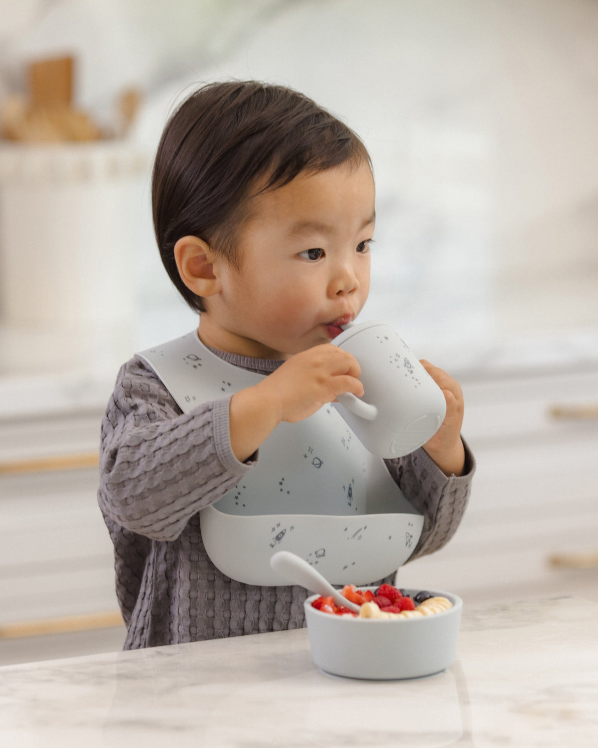 Toddler uses Rylee + Cru Feeding Kit Space, eating at the kitchen counter in a silicone bib.