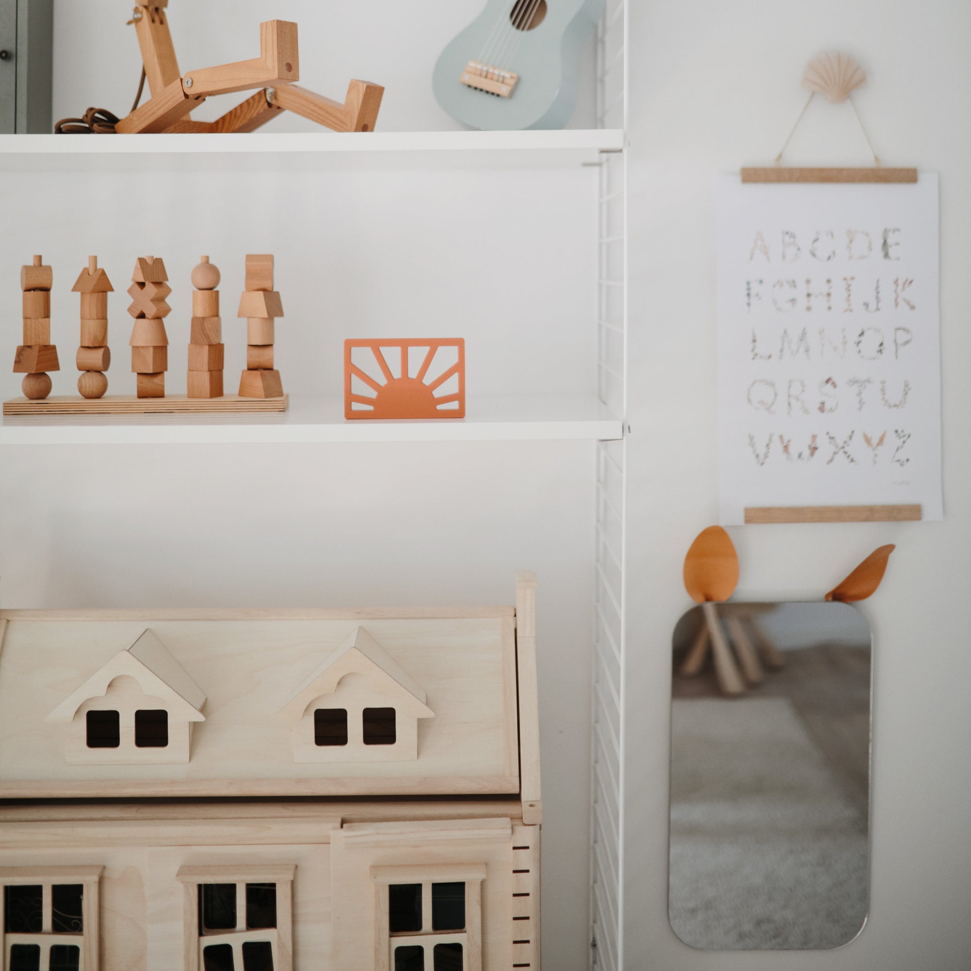A neatly organized children's room shelf featuring mushie's Sun Teether, wooden toys, a small dollhouse, a blue ukulele, an alphabet wall hanging, and a mirror with orange animal ears.