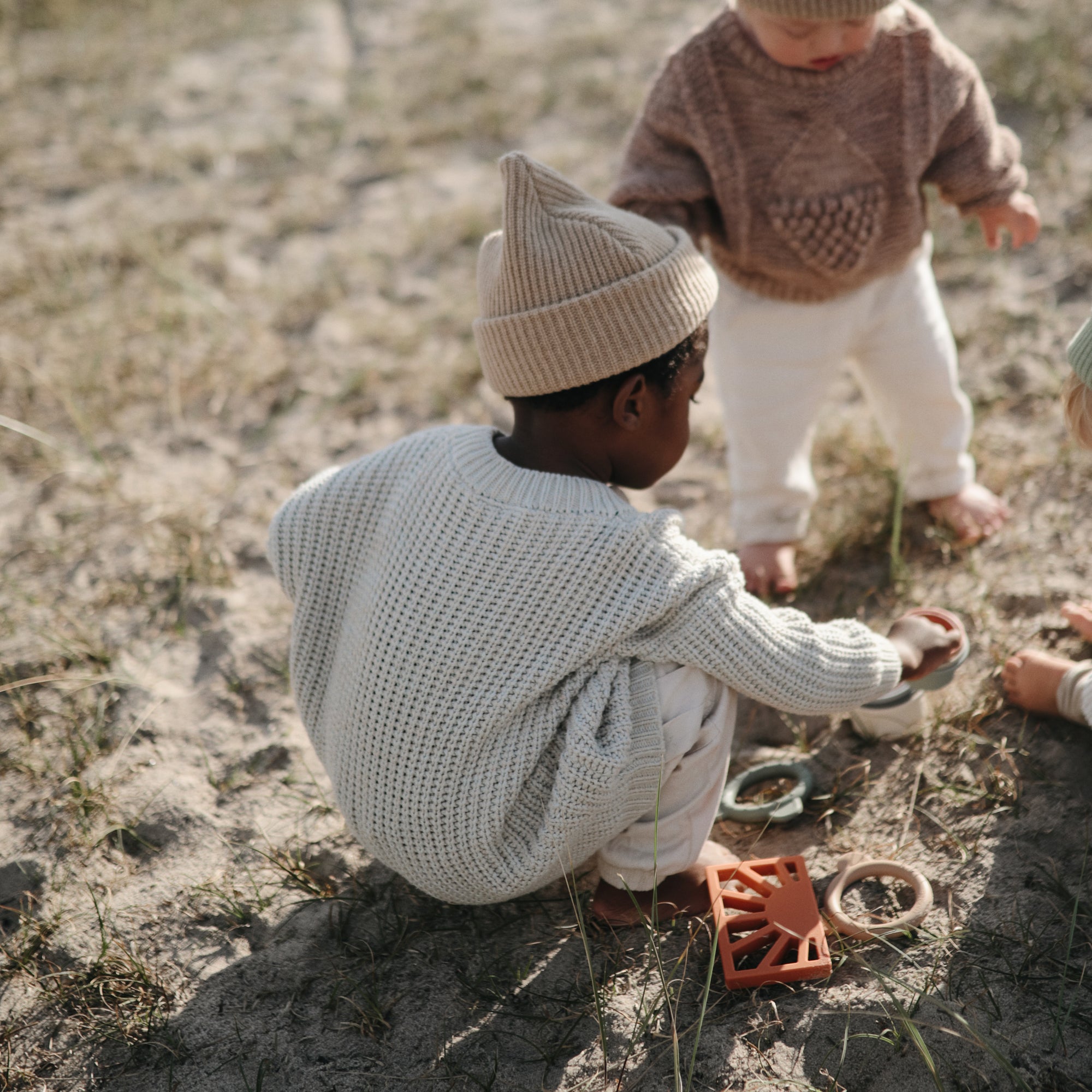 Two young children in knit sweaters and hats play outdoors on sandy ground with toys, including the mushie Sun Teether, surrounded by sparse grass.
