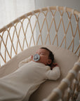 A baby in a cream-colored onesie sleeps in a light wooden bassinet fitted with the mushie Extra Soft Bassinet Muslin Crib Sheet, holding a pacifier as gentle daylight filters through the sheer curtain beside the crib.