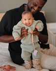A smiling baby in a green knit outfit and fuzzy boots is held by an adult on the bed. Nearby rests a FRIGG Butterfly Anatomical Silicone Pacifier 2-Pack (0-6 Months) from FRIGG, designed for baby's comfort.