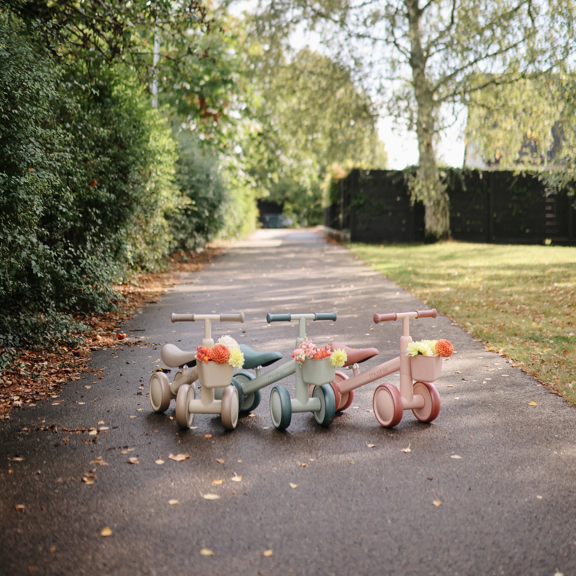 Three mushie Mini Toddler Bikes with flower baskets on a sunlit, tree-lined path.
