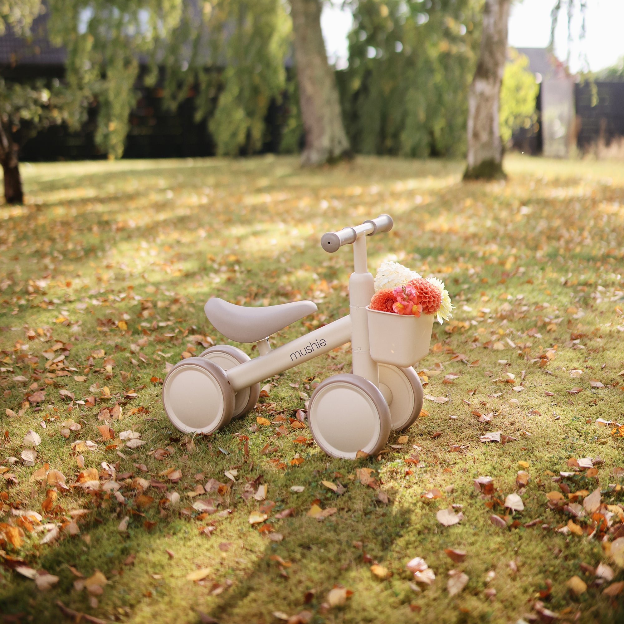 Mushie Mini Toddler Bike with floral basket on a grassy, leafy lawn outdoors.