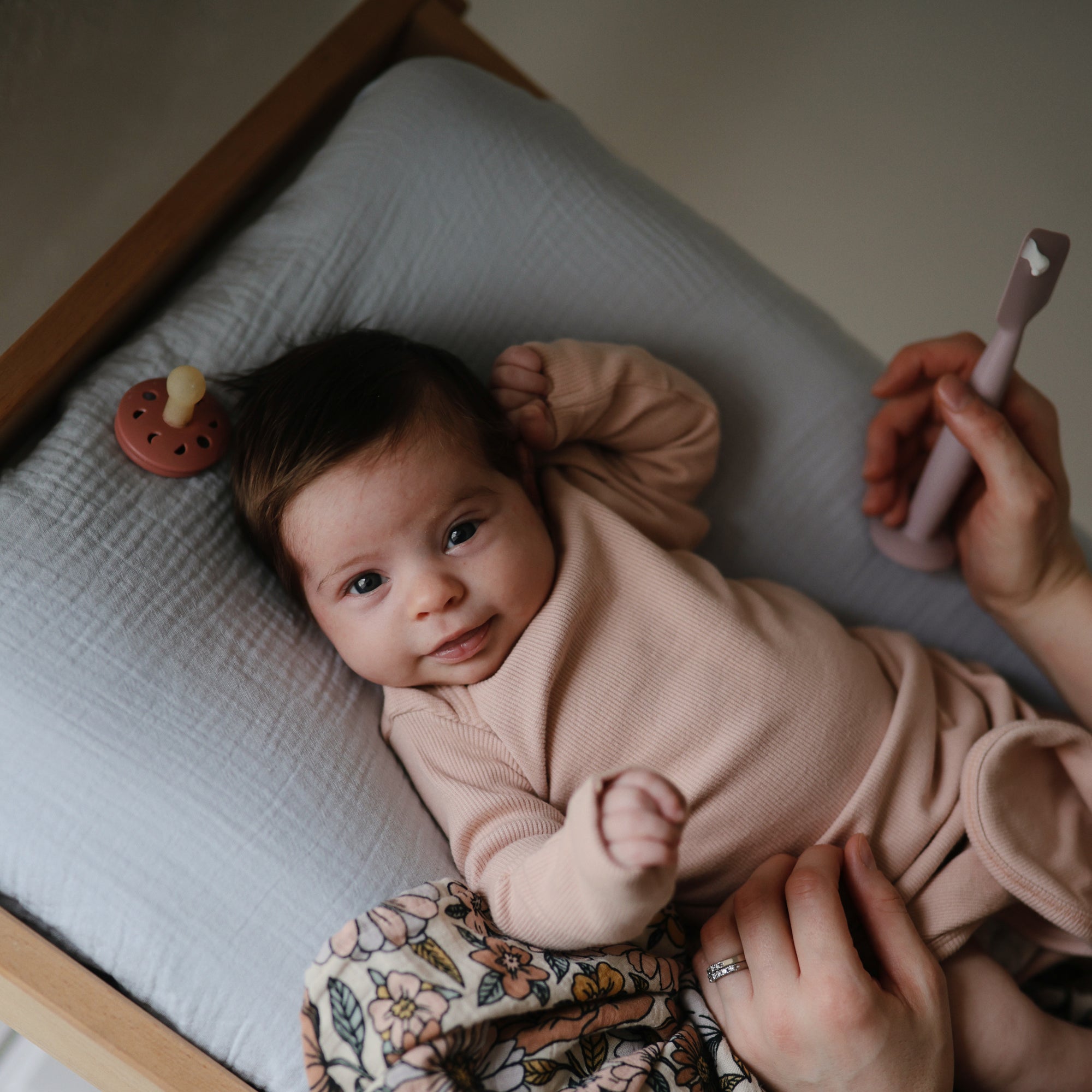A baby in a light pink outfit smiles on a cushioned changing table as an adult holds a pink toothbrush. Nearby, a FRIGG Moon Silicone Pacifier from the FRIGG 2-Pack rests among other baby essentials.