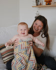 Smiling woman and baby on a bed, baby holding mushie's Figurine Teether and a blanket.