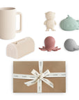 The mushie Sealife Bath Essentials Kit includes a ribbed beige mug, four silicone sea toys (bear in goggles, green submarine, pink octopus, grey whale), and a beige napkin holder, all packed in a brown gift box with a white ribbon.