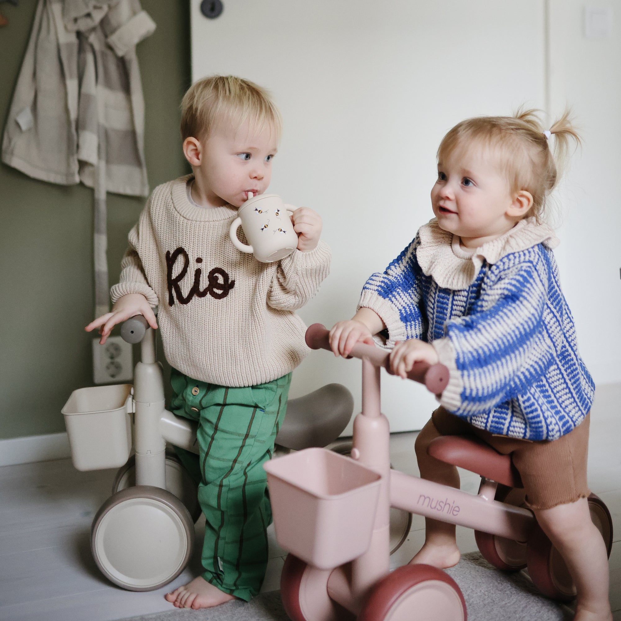 Two toddlers ride mushie Mini Toddler Bikes indoors; one drinks from a cup while the other watches.