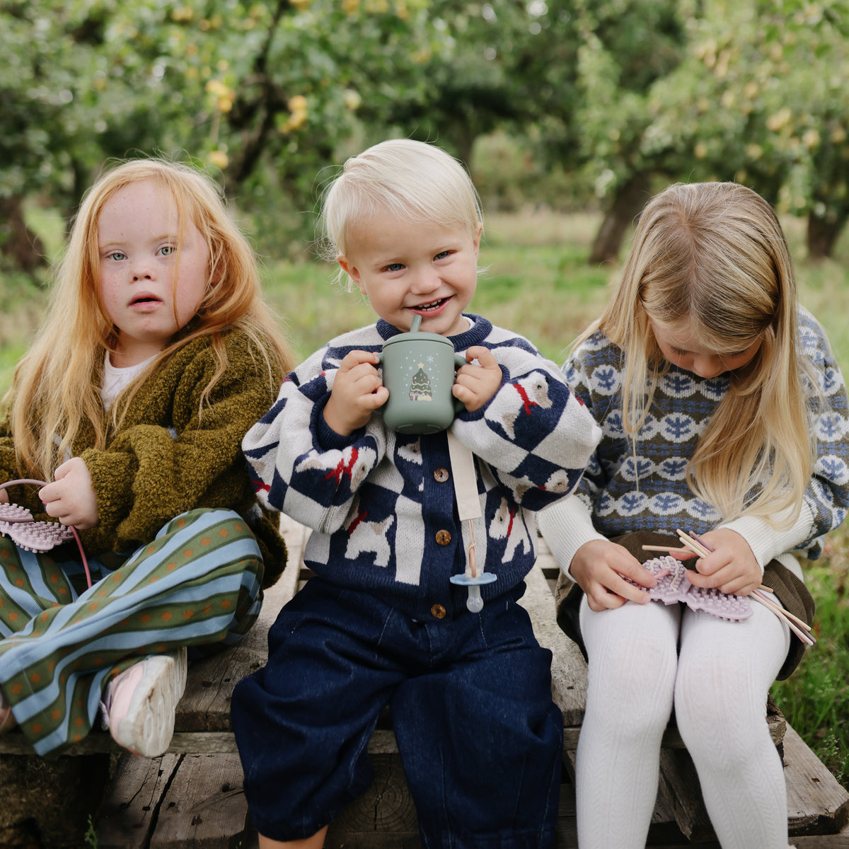 Three kids outdoors, two knitting, one with a Mushie Silicone Training Cup + Straw, all smiling.