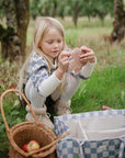 A girl weaves a heart on grass; her mushie Silicone Sensory Toy lies near apples and a basket.