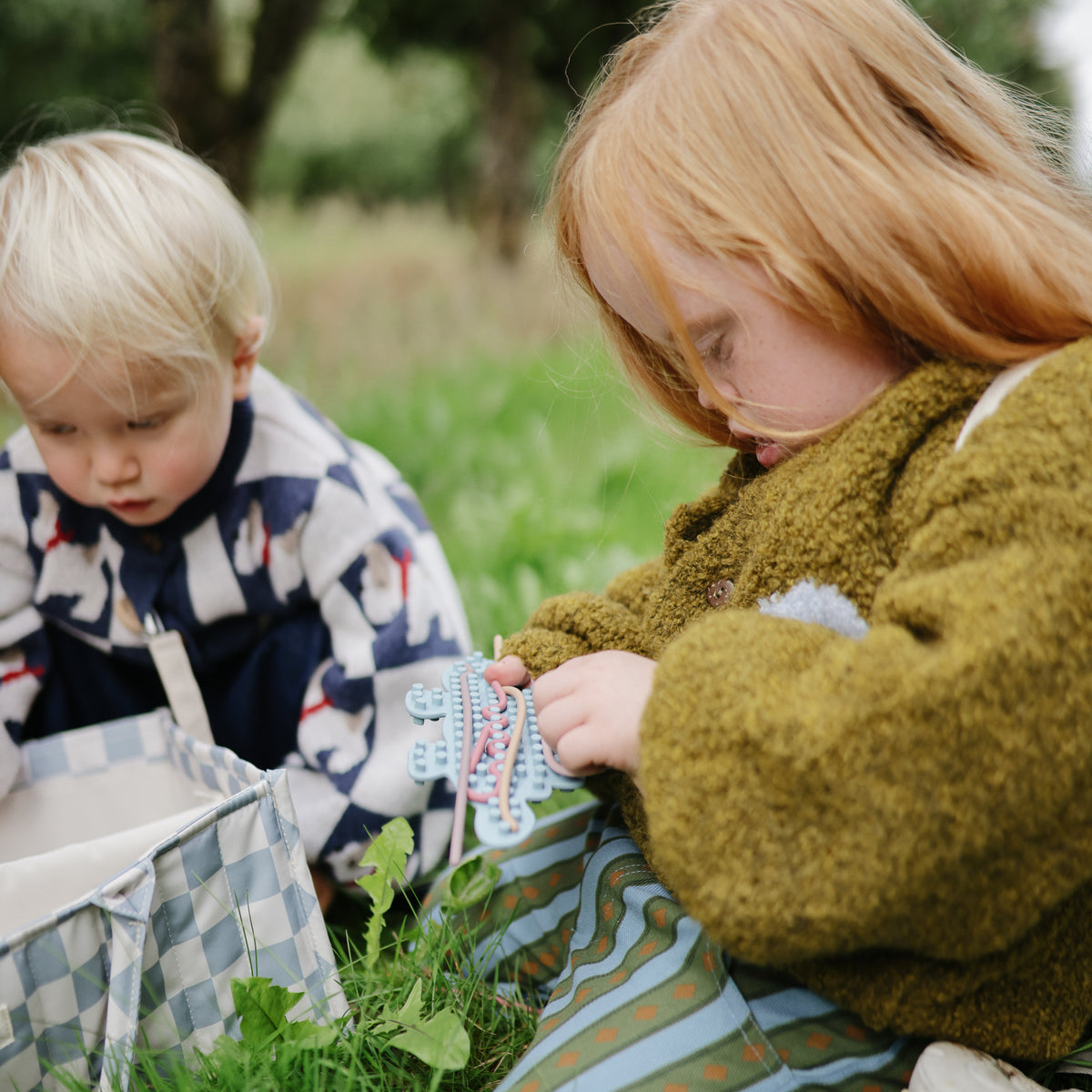 Two kids on grass, one playing with a mushie Silicone Sensory Toy in their hand.