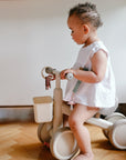 A toddler enjoys her first indoor ride on a beige mushie Mini Toddler Bike in a white dress.