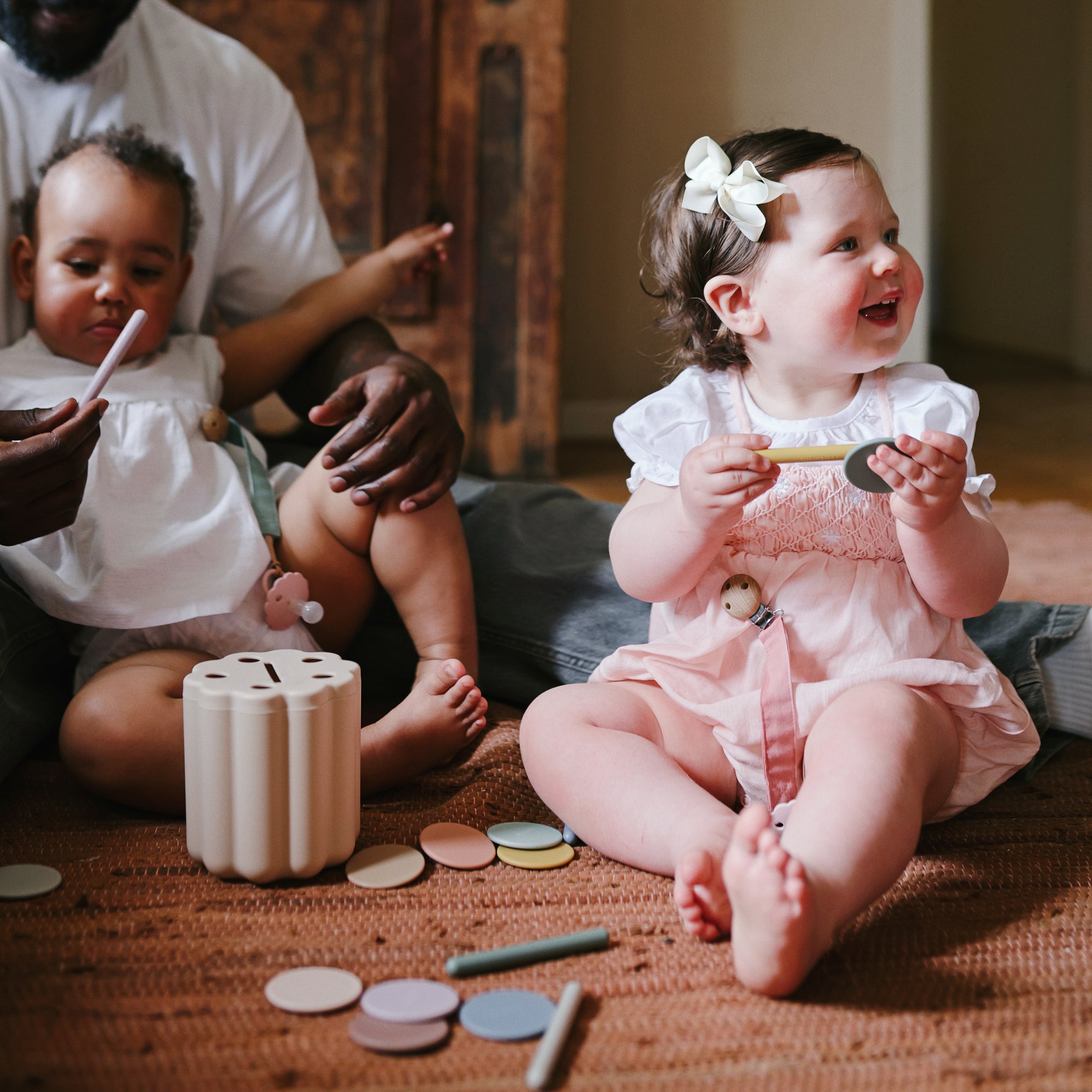 Two toddlers with wooden toys; one uses a mushie Linen Pacifier Clip 2-Pack. Adult in background.