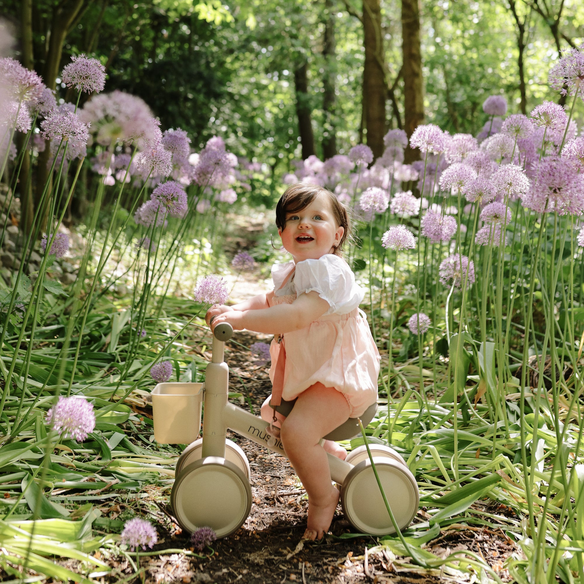 A smiling baby on a mushie Mini Toddler Bike rides among tall purple flowers on a sunny path.