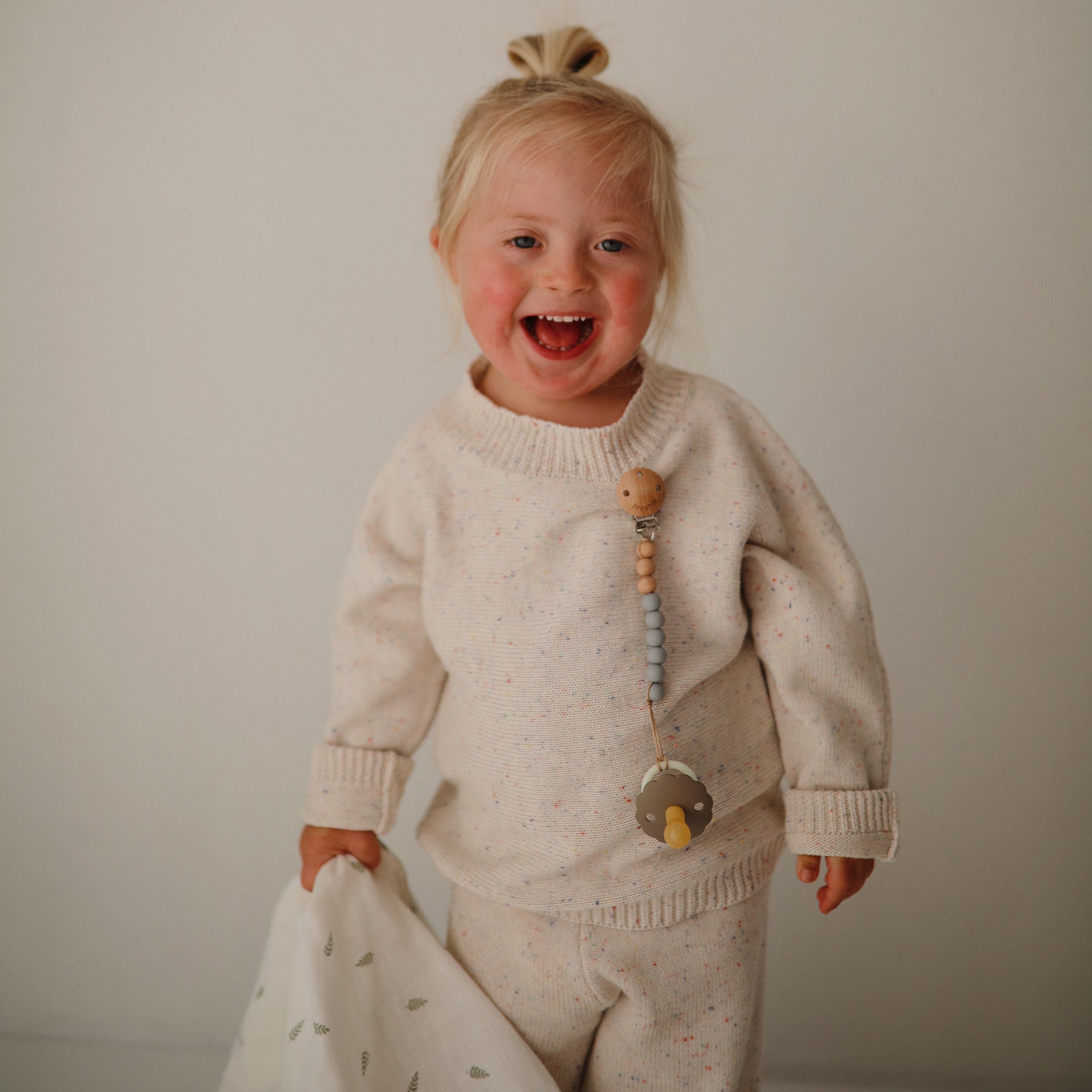 A smiling blonde child in a cream outfit holds a white blanket. A FRIGG Daisy Night Natural Rubber Pacifier from FRIGG, attached with a glow-in-the-dark wooden bead clip, is on their sweater. The background is plain and light-colored.