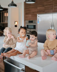 Four toddlers sit on a kitchen island with cups, cookies, and pacifiers. Nearby, FRIGG Andersen Fairytale Silicone Pacifier 2-Pack by FRIGG—made in Denmark—rests on the counter amid sleek appliances and wooden cabinets.