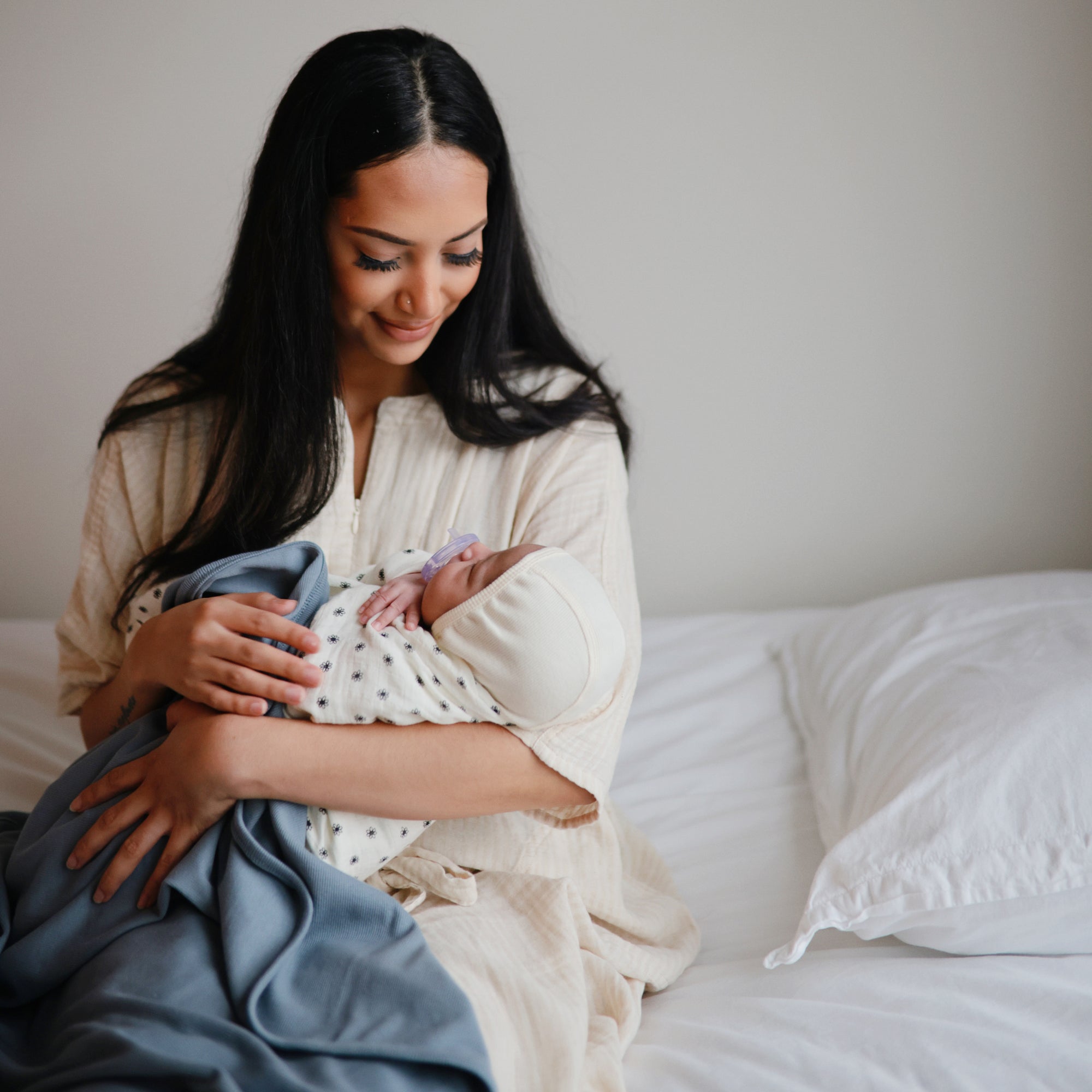 A woman in a light gown sits on a bed with white bedding, smiling at a baby cradled in her arms and wrapped in the Mushie Ribbed Baby Blanket. The room is softly lit, highlighting the blanket’s soft, breathable texture.