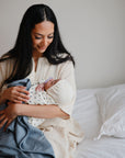 A woman in a light gown sits on a bed with white bedding, smiling at a baby cradled in her arms and wrapped in the Mushie Ribbed Baby Blanket. The room is softly lit, highlighting the blanket’s soft, breathable texture.