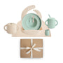 The mushie Deluxe Vanilla Confetti/Cambridge Blue Feeding Kit includes a speckled silicone bib, plate, spoon, fork, handled cup, sippy cup, and small container—all toddler feeding essentials in soft pastels presented as a perfect gift.
