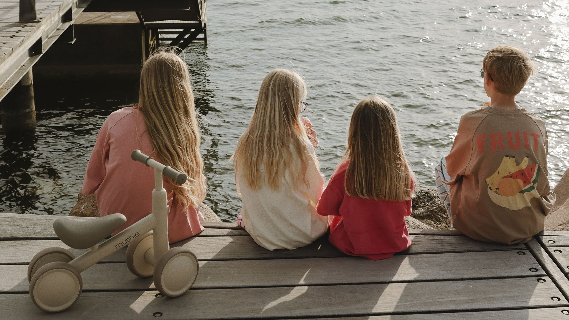 Children sit side by side on the edge of a wooden boardwalk, looking out over calm water on a peaceful day.