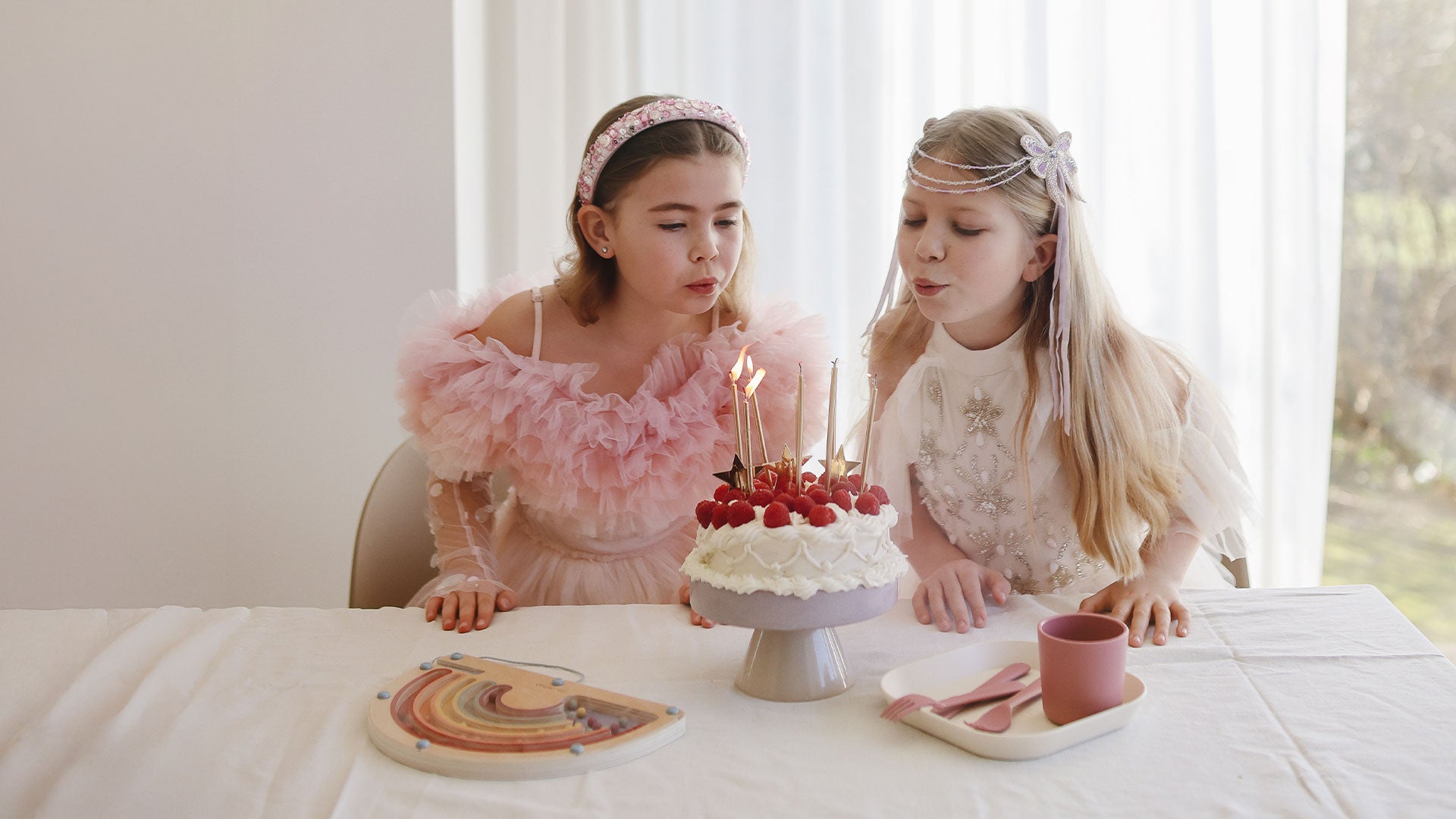 Two children celebrate a birthday by blowing out candles on a cake. A Mushie Rainbow Maze and feeding essentials are displayed on the table alongside the birthday cake.