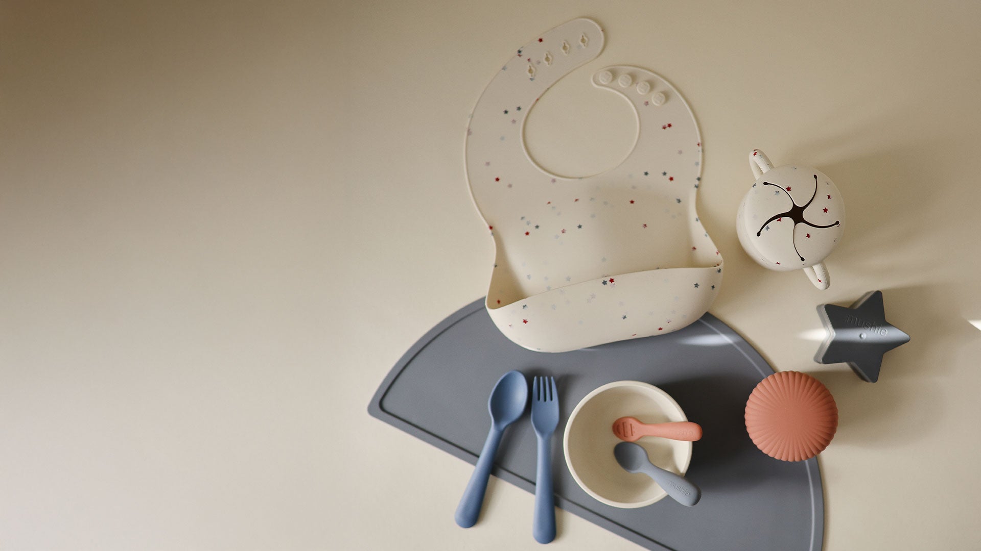 New Mushie silicone bib and silicone snack cup featuring the limited edition Star Confetti print with scattered mushie feeding products on a soft neutral background.