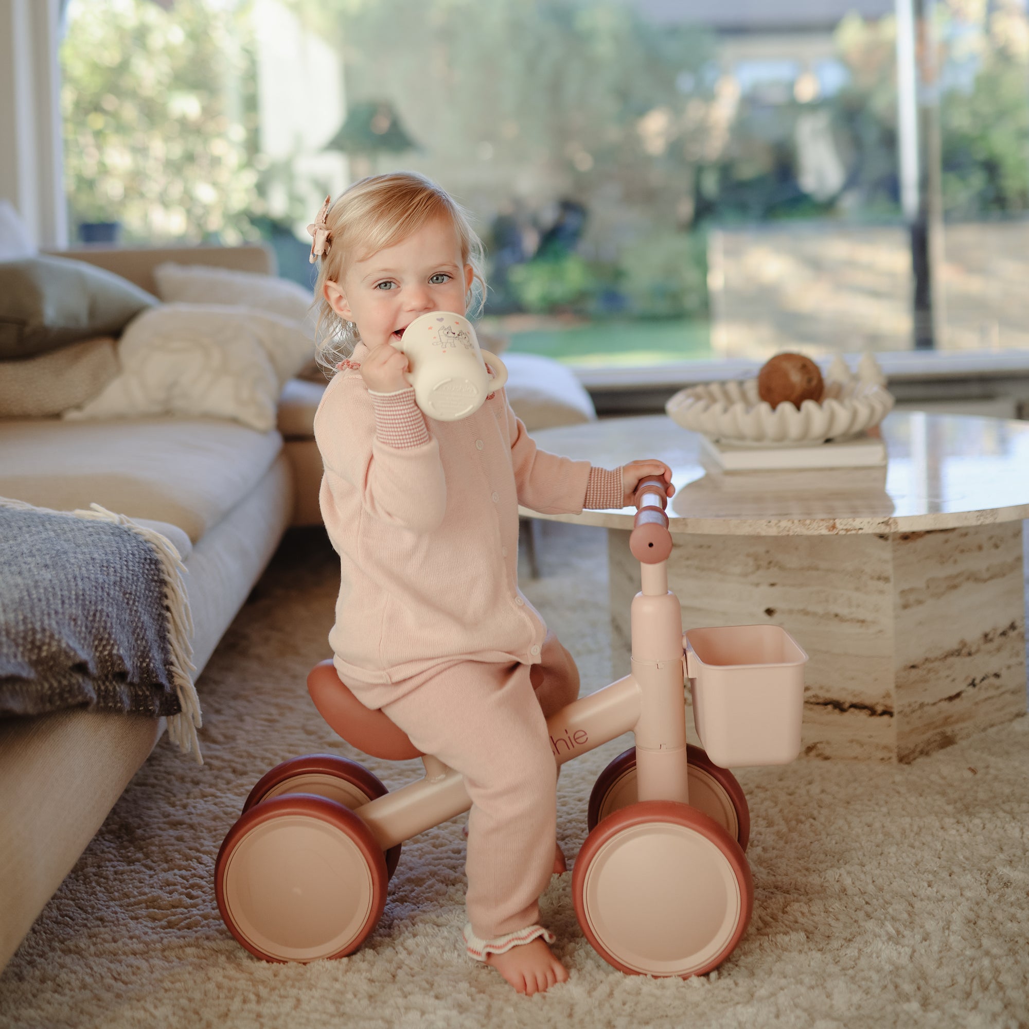 Toddler in pink pajamas rides mushie Mini Toddler Bike indoors while drinking from a bottle.