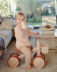 Toddler in pink pajamas rides mushie Mini Toddler Bike indoors while drinking from a bottle.