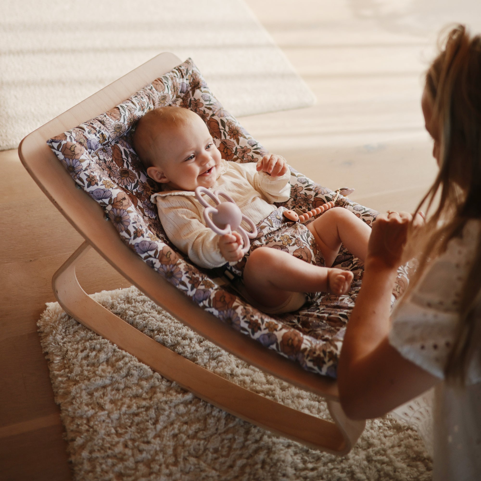 Smiling baby holds a mushie Daisy Rattle Teether while relaxing in a patterned rocker.