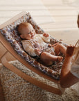 Smiling baby holds a mushie Daisy Rattle Teether while relaxing in a patterned rocker.