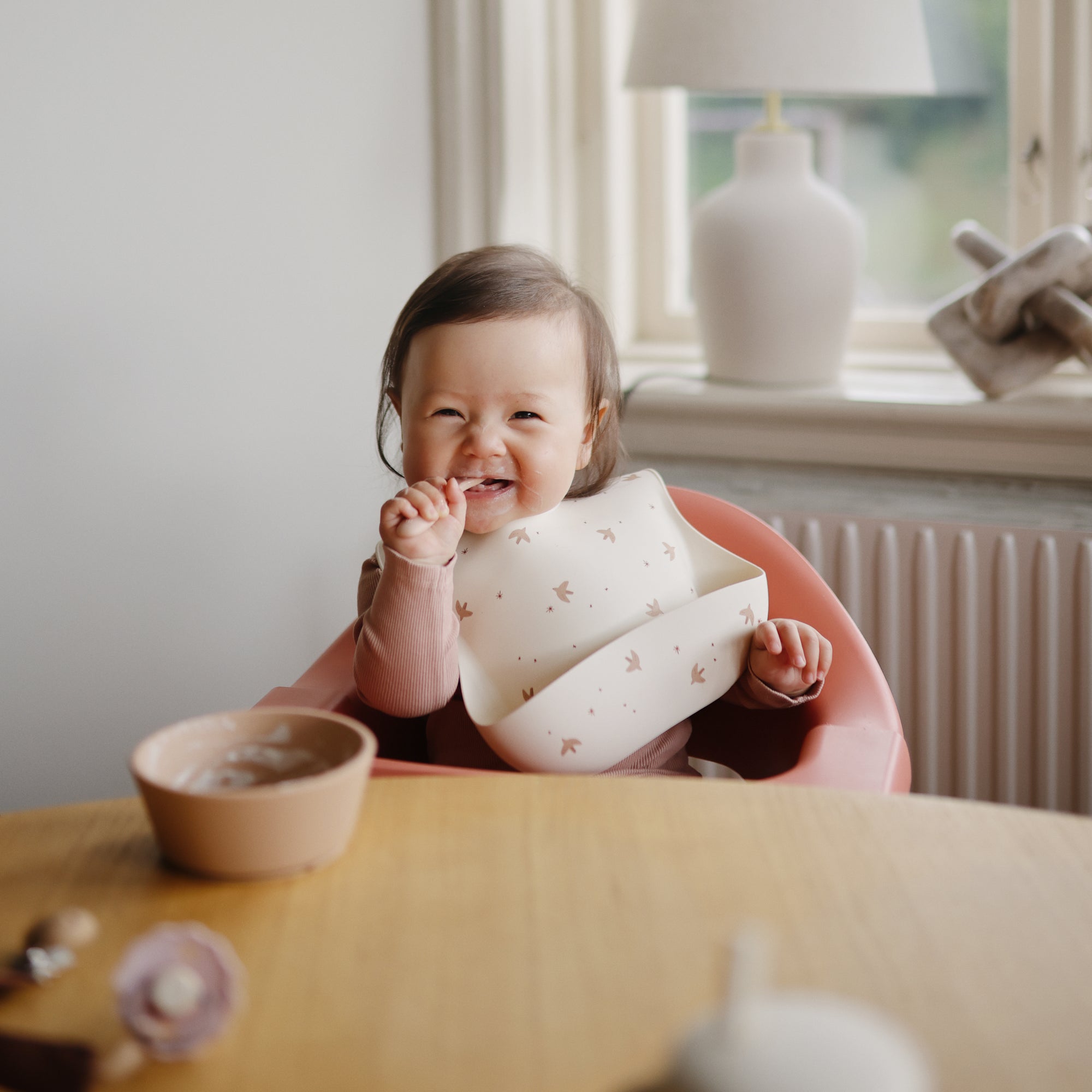 Smiling baby wears mushie Silicone Baby Bib in a highchair, holding food by a bowl.
