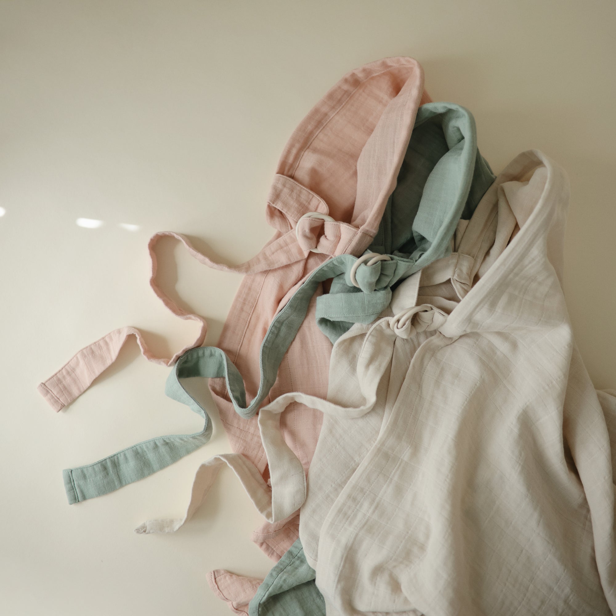 The mushie Muslin Nursing Cover, shown in peach, sage green, and off-white, features soft muslin fabric with adjustable straps, highlighting its versatility as a multi-use nursing cover.