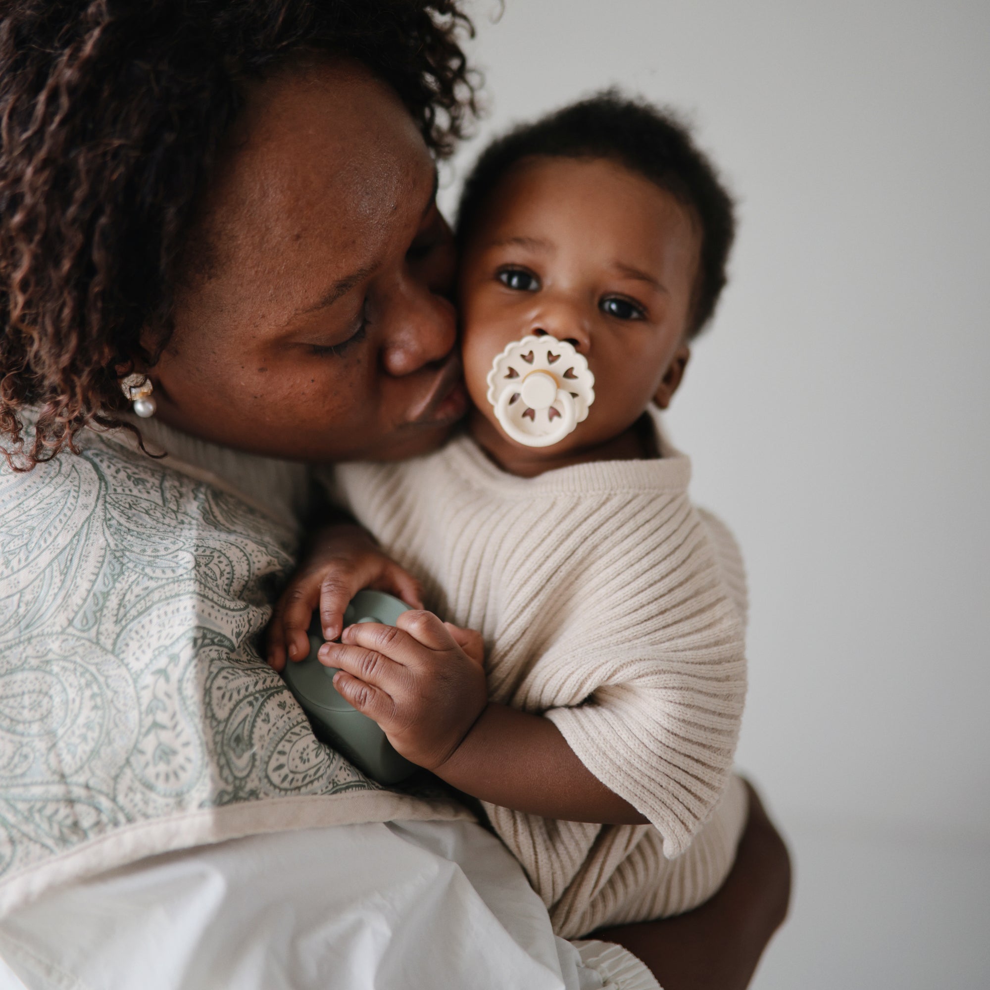A woman lovingly kisses a baby on the cheek. The baby, wearing beige and holding a rattle, looks at the camera with a FRIGG Andersen Fairytale Silicone Pacifier from FRIGG in their mouth. The background is softly blurred.