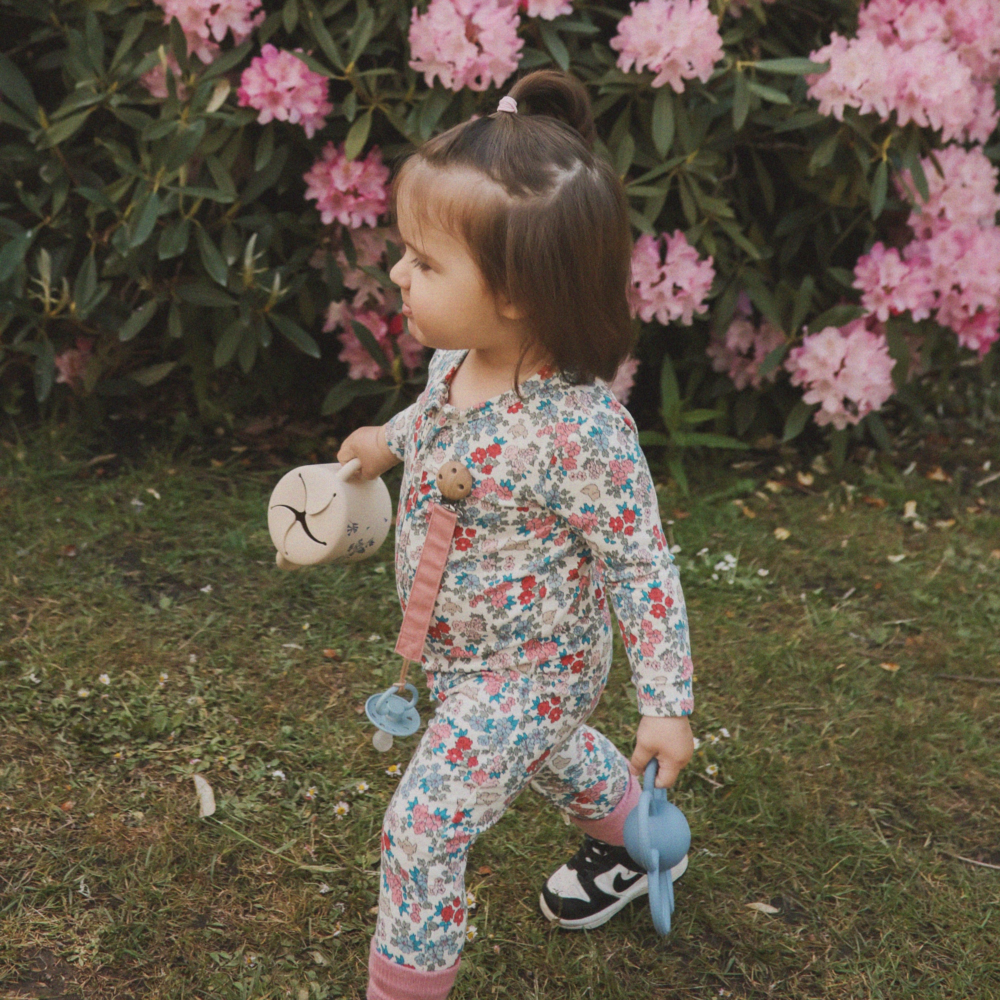 A young child in a floral outfit walks on grass holding a blue sippy cup, a beige bottle, and the mushie Linen Pacifier Clip 2-Pack, with pink rhododendron flowers and green leaves in the background.