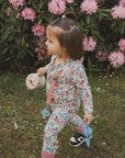 A young child in a floral outfit walks on grass holding a blue sippy cup, a beige bottle, and the mushie Linen Pacifier Clip 2-Pack, with pink rhododendron flowers and green leaves in the background.