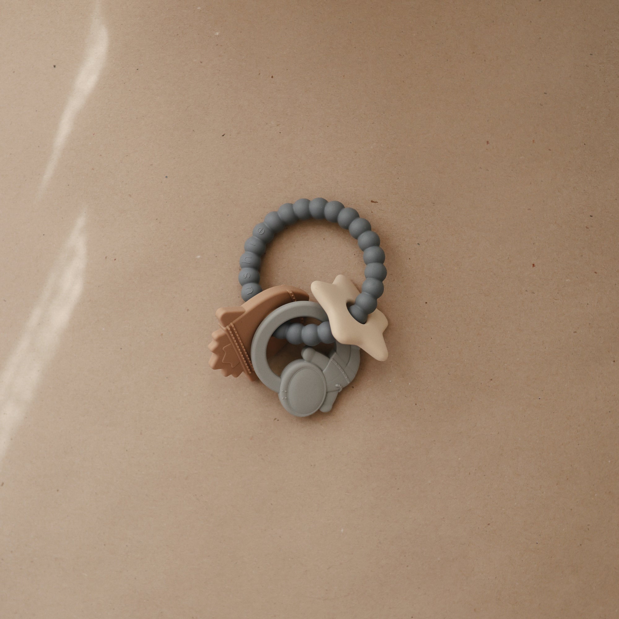 A silicone baby teething ring with beaded texture and attached star, sun, and moon-shaped charms rests on a light brown surface.