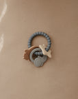 A silicone baby teething ring with beaded texture and attached star, sun, and moon-shaped charms rests on a light brown surface.