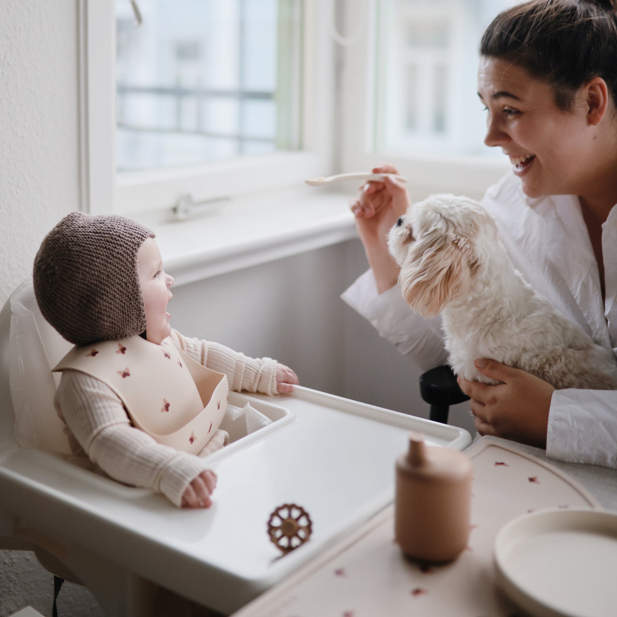 A woman smiles while feeding a baby in a high chair. The baby, wearing a bib and knit bonnet, enjoys a FRIGG Andersen Fairytale Natural Rubber Pacifier by the window, with a small white dog on the woman's lap.