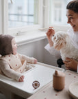 A woman smiles while feeding a baby in a high chair. The baby, wearing a bib and knit bonnet, enjoys a FRIGG Andersen Fairytale Natural Rubber Pacifier by the window, with a small white dog on the woman's lap.