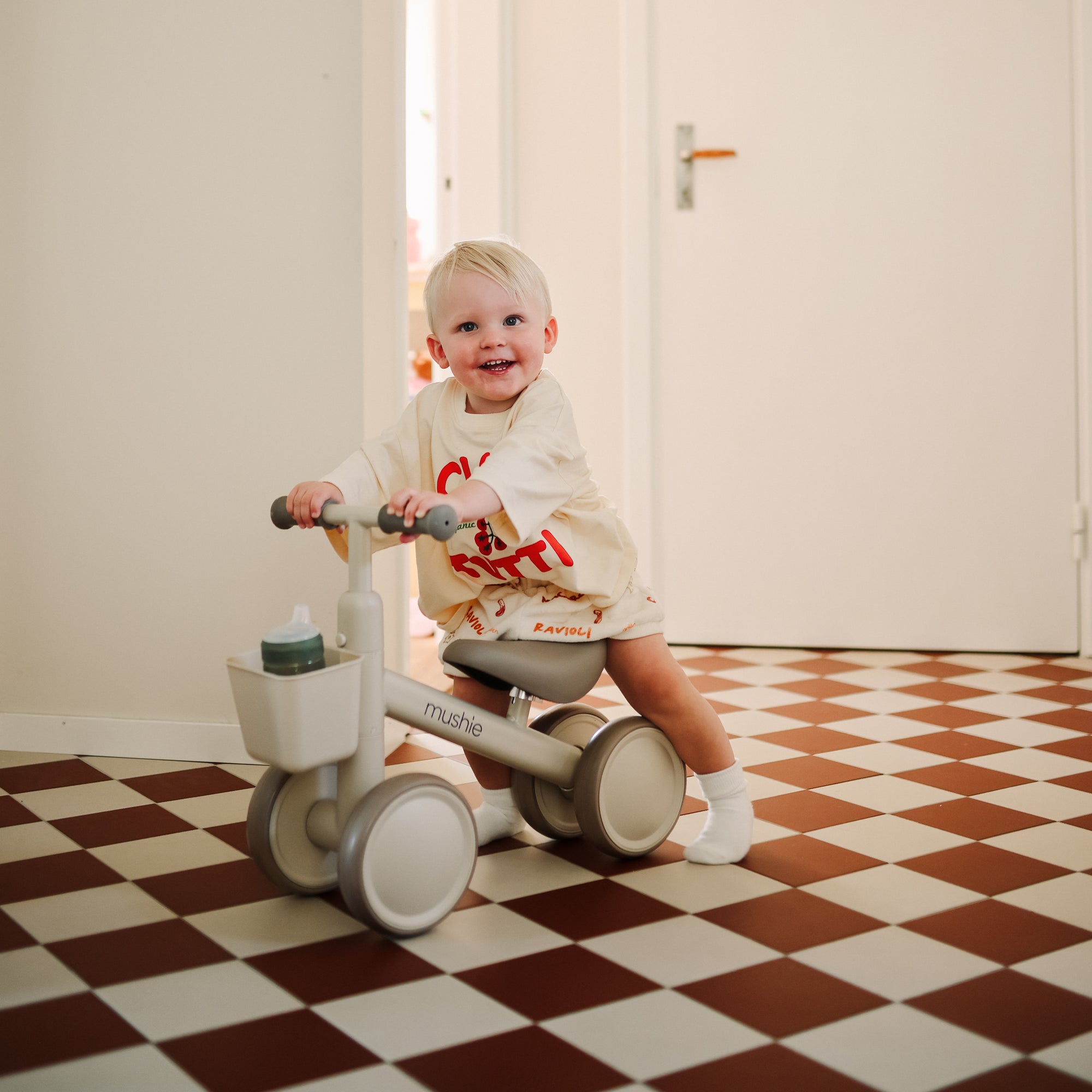A smiling toddler rides the mushie Mini Toddler Bike on a red and white checkered floor.