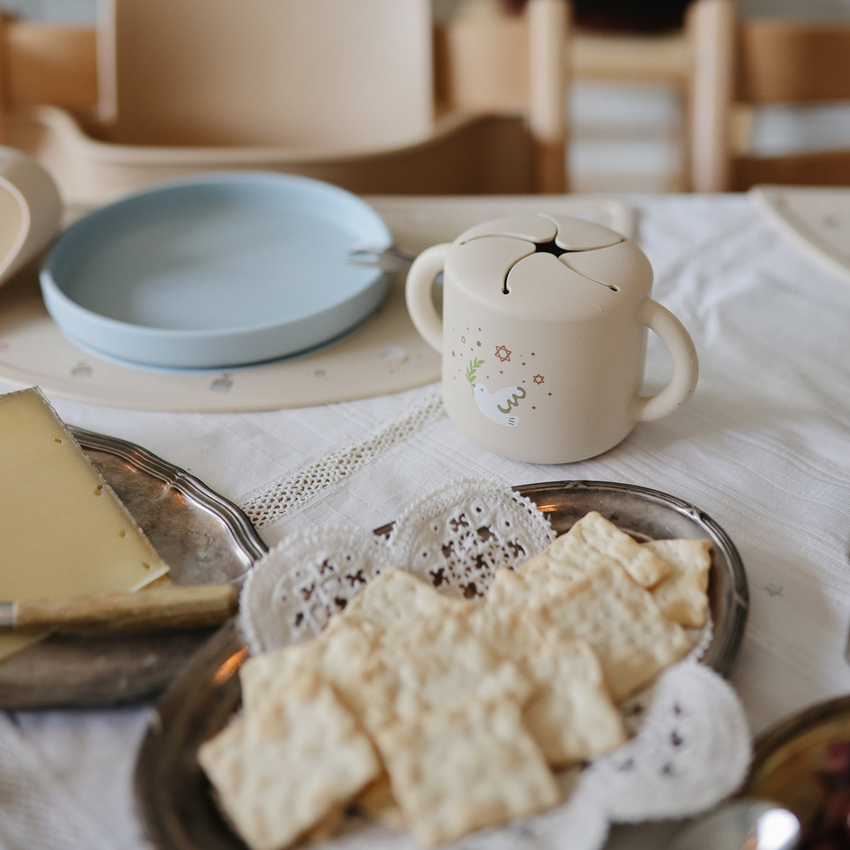 A table with crackers, cheese, a blue plate, and a mushie Snack Cup with handles.