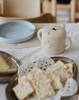 A table with crackers, cheese, a blue plate, and a mushie Snack Cup with handles.
