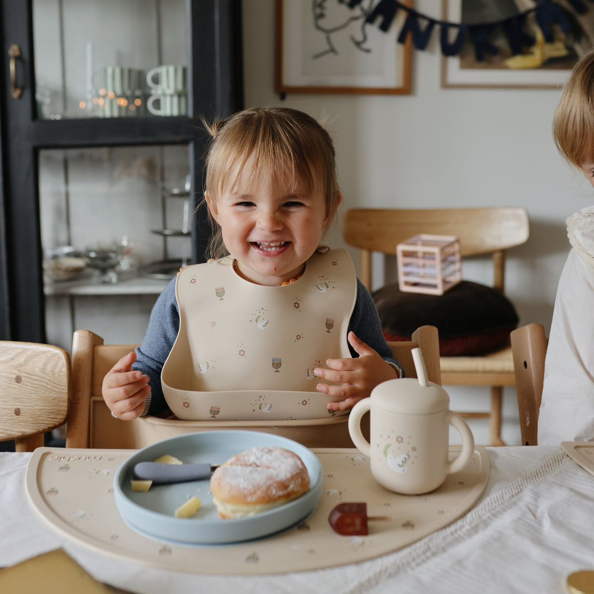 Toddler in mushie Silicone Baby Bib sits at table with food, donut, and a cup.