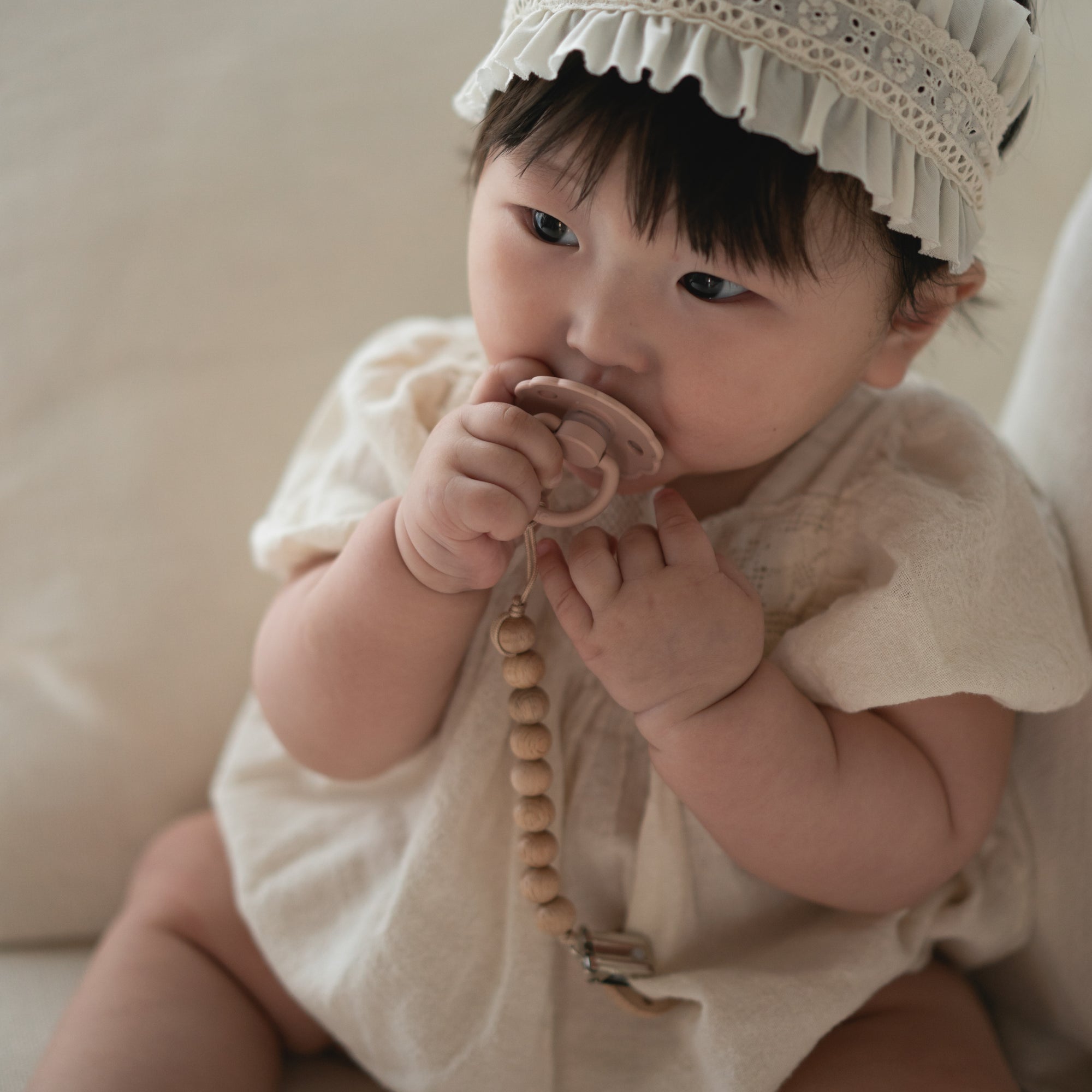 Baby in white with headband holds FRIGG Daisy Natural Rubber Pacifier 2-Pack with beaded clip.