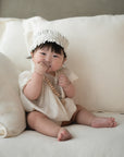 Baby in white holds FRIGG Daisy Natural Rubber Pacifier 2-Pack on a cream sofa.