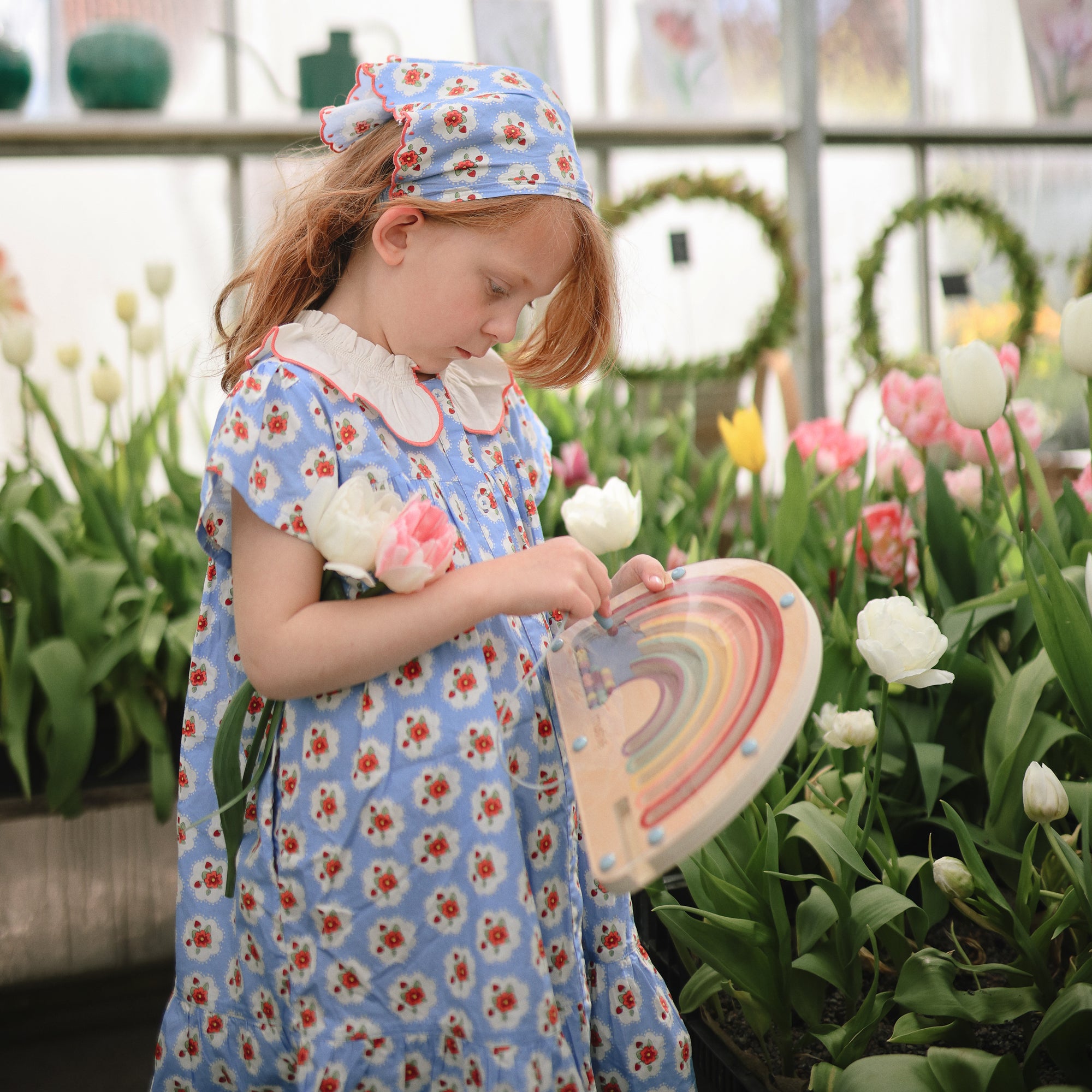 Young girl in a blue floral dress holds the mushie Wooden Magnetic Rainbow Maze by tulips.