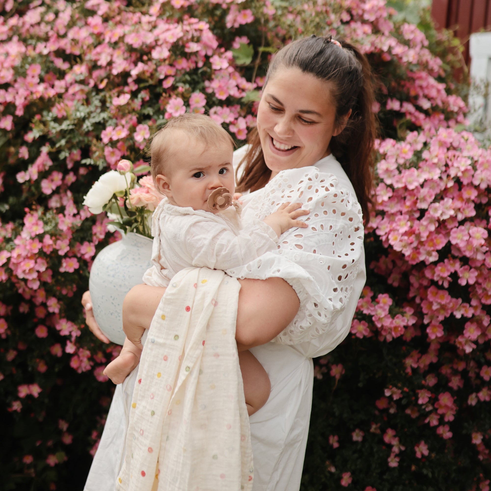 Woman holds baby with FRIGG Daisy Natural Rubber Pacifier near blooming pink flowers and a vase.